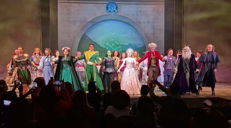 Elenco do musical Wicked de mãos dadas no palco, acenando e agradecendo ao público sob luzes coloridas.