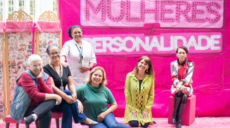 Foto mostra seis mulheres - cinco delas sentadas e uma em pé - posando na quadra do centro de atendimento em frente a um painel cor de rosa montado para a ação, em que se lê Mulheres de Personalidade