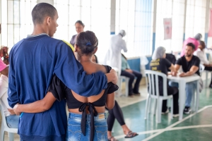Foto mostra os diversos atendimentos na quadra ensolarada do centro socioeducativo. Ao fundo, há pessoas sentadas e em pé. Em primeiro plano, aparecem do lado esquerdo um adolescente abraçado, de costas, para uma mulher, sem identificá-los