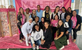 Imagem mostra 17 mulheres com cabelos trançados que participaram da ação. Elas estão na quadra do centro de atendimento, posando num cenário montado: biombo e tecidos na cor rosa, escrito no tecido Mulheres de Personalidade