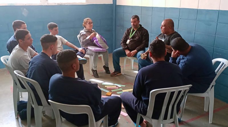 Palavras como “Ética”, “Amor” e “Justiça” estão dispostas no chão, junto de livros e objetos, enquanto jovens com chinelos participam da dinâmica.