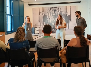 Foto mostra, em primeiro plano, três adultos sentados em cadeiras, numa das salas do museu. Eles observam três adultos em pé - duas mulheres e um homem -, sendo que uma das mulheres fala e gesticula. Atrás deles já uma foto em preto e branco