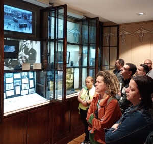 Foto mostra ao menos sete adultos em pé, entre homens e mulheres,, em uma das dependências do museu. Eles observam diversas fotos do acervo de Mário de Andrade numa parece com decoração em madeira e janelas.