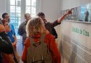 Foto mostra seis adultos em uma das dependências do museu. Um deles aponta para fotos dispostas numa parede branca, com conteúdo escrito abaixo, quanto os outros cinco adultos observam