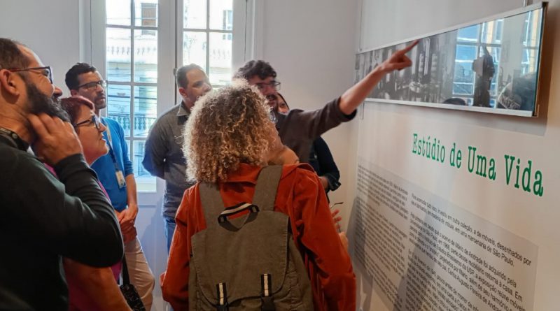 Foto mostra seis adultos em uma das dependências do museu. Um deles aponta para fotos dispostas numa parede branca, com conteúdo escrito abaixo, quanto os outros cinco adultos observam