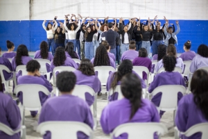 Foto mostra a apresentação do coral na quadra coberta do centro socioeducativo. De costas para quem observa a imagem, há 22 adolescentes sentadas em cadeiras e vestindo uniforme na cor lilás. Ao fundo, no palco, está o coral, formado por jovens de ambos os sexos, cantando e gesticulando com os braços para cima, sob regência do maestro.