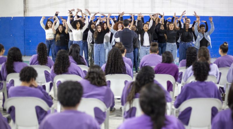 Foto mostra a apresentação do coral na quadra coberta do centro socioeducativo. De costas para quem observa a imagem, há 22 adolescentes sentadas em cadeiras e vestindo uniforme na cor lilás. Ao fundo, no palco, está o coral, formado por jovens de ambos os sexos, cantando e gesticulando com os braços para cima, sob regência do maestro.
