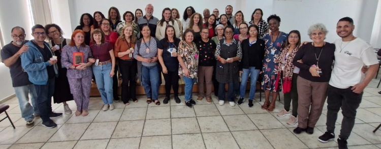 Foto mostra um grupo de 36 pessoas, entre mulheres e homens, em pé, próximo ao palco do auditório do Complexo do Brás. As pessoas estão próximas umas às outras, em foto posada do grupo. A maioria sorri.