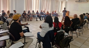 Foto mostra o auditório do Complexo, com paredes brancas e persianas ao fundo. Em formato de roda, estão homens e mulheres adultos sentados em carteiras tipo universitárias. Ao centro da roda está a palestrante, uma mulher negra. Atrás dela está projetada uma apresentação na parede.