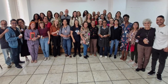 Foto mostra um grupo de 36 pessoas, entre mulheres e homens, em pé, próximo ao palco do auditório do Complexo do Brás. As pessoas estão próximas umas às outras, em foto posada do grupo. A maioria sorri.