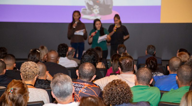 Três adolescentes, todas meninas, falam ao microfone no palco diante de uma plateia atenta no auditório do Museu da Língua Portuguesa.