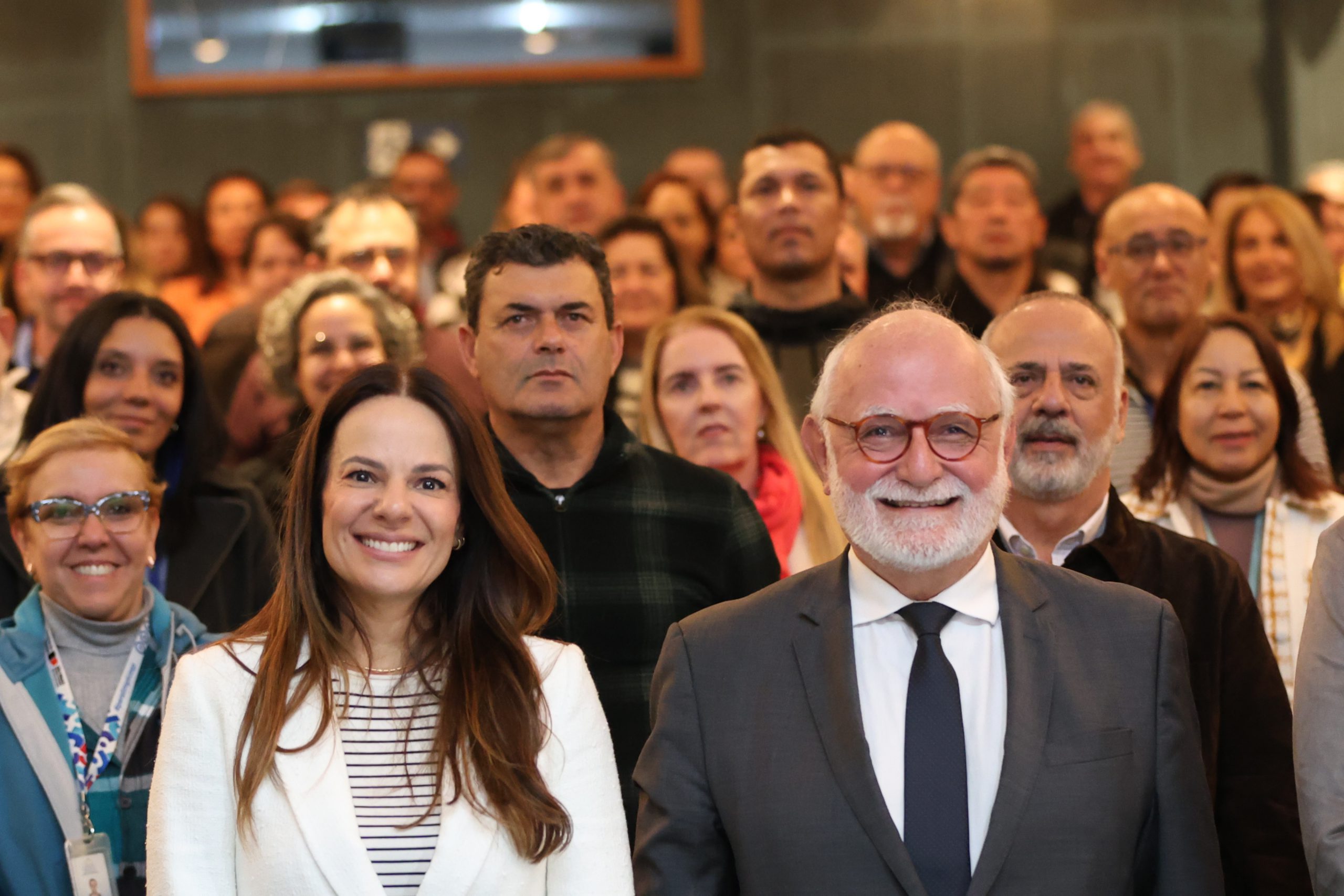 A imagem mostra um grupo de pessoas em um ambiente interno, que parece ser um auditório ou sala de eventos, com a plateia ao fundo e duas pessoas em destaque em primeiro plano.  Em primeiro plano, uma mulher e um homem sorriem para a câmera. A mulher, à esquerda, tem cabelos longos e castanhos, veste um blazer branco e uma blusa listrada por baixo. Ao lado dela, à direita, o homem tem barba e cabelos grisalhos, usa óculos, um terno cinza-escuro e uma gravata preta.  Atrás deles, um grupo diversificado de pessoas, homens e mulheres de várias idades e etnias, compõe a plateia, alguns com expressões sorridentes, outros mais neutras. O foco principal está nas duas figuras em primeiro plano, enquanto as pessoas ao fundo estão ligeiramente desfocadas. A iluminação geral parece ser de ambiente, com um tom natural.