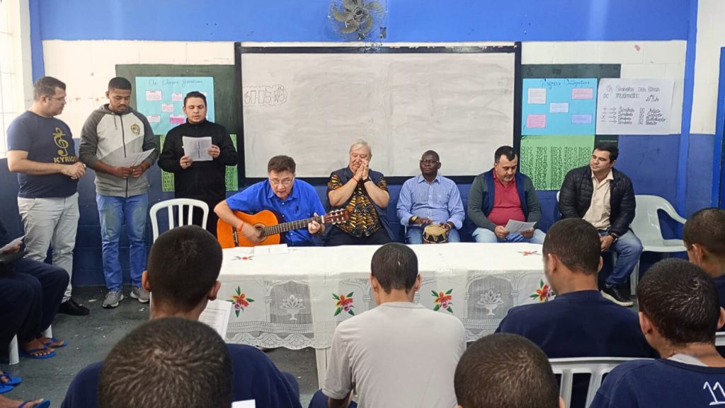 Grupo da Fazenda da Esperança canta e toca instrumentos para adolescentes sentados em uma sala no CASA.