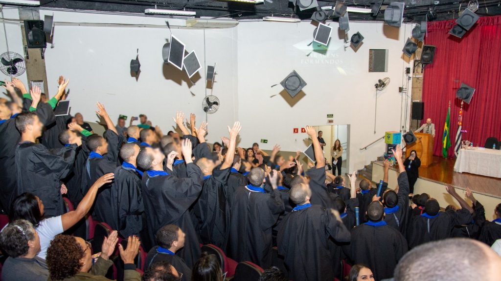 Adolescentes com becas comemoram formatura jogando seus capelos para o alto em auditório.