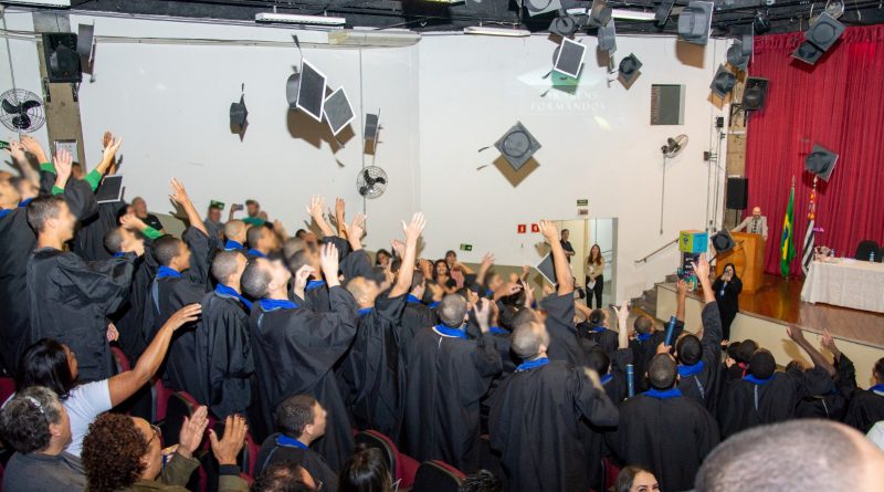 Adolescentes com becas comemoram formatura jogando seus capelos para o alto em auditório.