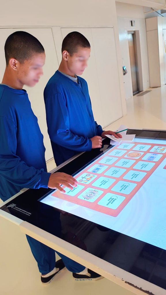 Dois adolescentes utilizam mesa interativa digital com imagens e textos sobre moedas e símbolos nacionais.
