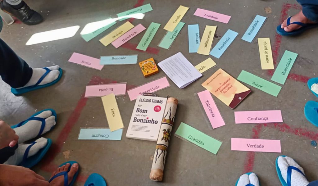 Palavras como “Ética”, “Amor” e “Justiça” estão dispostas no chão, junto de livros e objetos, enquanto jovens com chinelos participam da dinâmica.
