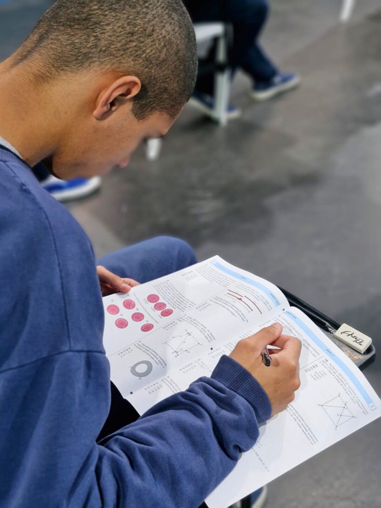 Adolescente segura caneta e responde prova da OBMEP com questões ilustradas em um caderno.