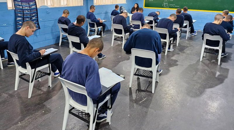 Jovens uniformizados sentados em sala de aula fazendo prova escrita, com parede azul ao fundo e lousa verde.