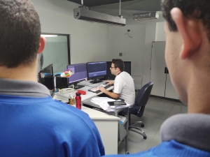 Foto mostra uma sala fechada, com paredes brancas, em que há um vridro em uma delas. Há uma mesa grande com quatro monitores de computador. Sentado à mesa, está um adulto, o editor da novela A Caverna Encantada, demonstrando como é feita a edição da novela. Em primeiro plano, aparecem dois adolescentes, de costas, observando a edição