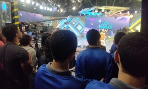 Foto mostra o estúdio onde está sendo gravado o programa Domingo Legal. Ao fundo, aparece a logomarca colorida em azul do programa, tendo à frente o apresentador Celso Portiolli comandando a brincadeira de torta na cara: duas fãs respondem sobre a cantora Joelma, convidada. Em primeiro plano aparecem três dos jovens do centro de atendimento de costas, observando a gravação