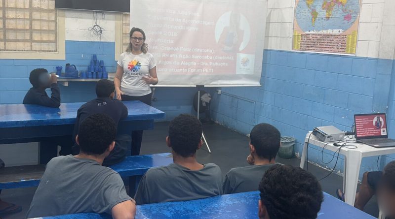 Fotografia mostra adolescentes, sentados em um refeitório com mesas de alvenaria, assistindo a uma palestra sobre empregabilidade. Os jovens estão todos de costas, usando camisetas cinzas e prestando atenção à fala da palestrante.