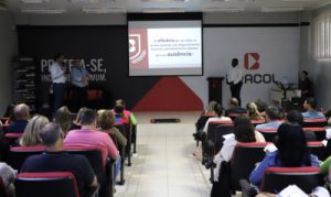 Foto mostra o auditório da Bracol. Ao fundo há um painel com a logomarca e o slogan da empresa, tendo à sua frente uma tela de projeção e um palco. Nesse palco há três pessoas: duas do lado esquerdo e uma do lado direito. Na plateia há cadeiras estofadas totalmente ocupadas por adultos sentados