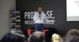 Foto mostra o palestrante em cima do palco, falando e gesticulando para a plateia sentada em cadeiras estofadas. Atrás do palestrante há um painel preto com o slogan e produtos da empresa