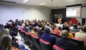 Foto mostra o auditório da Bracol, uma sala ampla, com cerca de 80 cadeiras estofadas, ocupadas por adultos sentados. Ao fundo, um homem recepciona os servidores, em pé no palco, tendo ao fundo um painel preto com o slogan e produtos da empresa