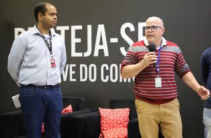 Foto mostra em detalhe dois adultos em pé no palco do auditório: o palestrante, um homem negro do lado esquerdo, e um homem branco, segurando microfone e falando, do lado direito. Ao fundo, aparece o painel com o slogan da empresa