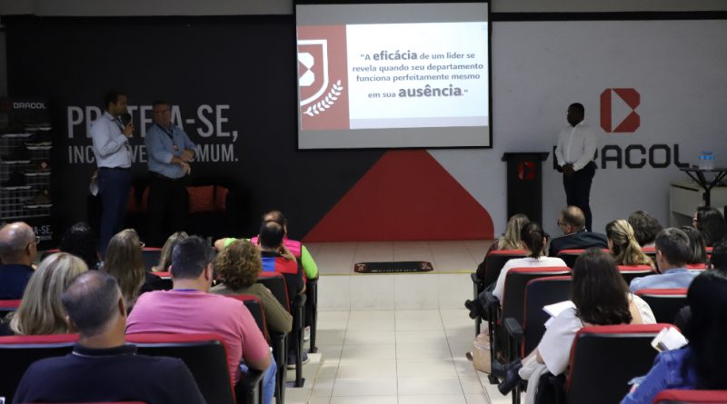 Foto mostra o auditório da Bracol. Ao fundo há um painel com a logomarca e o slogan da empresa, tendo à sua frente uma tela de projeção e um palco. Nesse palco há três pessoas: duas do lado esquerdo e uma do lado direito. Na plateia há cadeiras estofadas totalmente ocupadas por adultos sentados