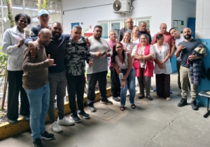 Foto mostra 17 servidores na entrada da área técnica do CASA Peruíbe, onde há um pequeno jardim e, ao fundo, grade de acesso e parede com janela e porta. São homens e mulheres que estão posicionados um ao lado do outro, posando para a foto