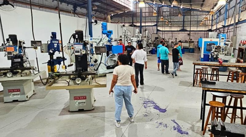 Adolescentes e equipe caminham por entre máquinas e bancadas em um amplo galpão industrial do IFSP.