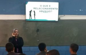 Fotografia retrata uma mulher, loira de pele branca, que é representante do projeto falando sobre a questão. Ao fundo está uma lousa, com uma televisão exibindo uma apresentação. Na parte de baixo, estão adolescentes assistindo à palestra.