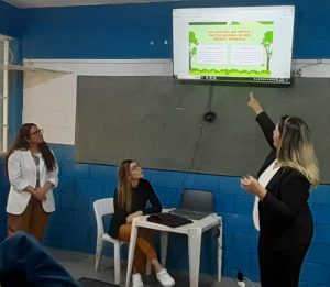Fotografia ilustra momento da palestra. Na foto, estão três mulheres, a direita, uma delas aponta para uma televisão onde é mostrada uma apresentação. No centro, está uma mulher mais jovem, sentada passando os slides. Na esquerda, está a teceria mulher, que também gesticula.