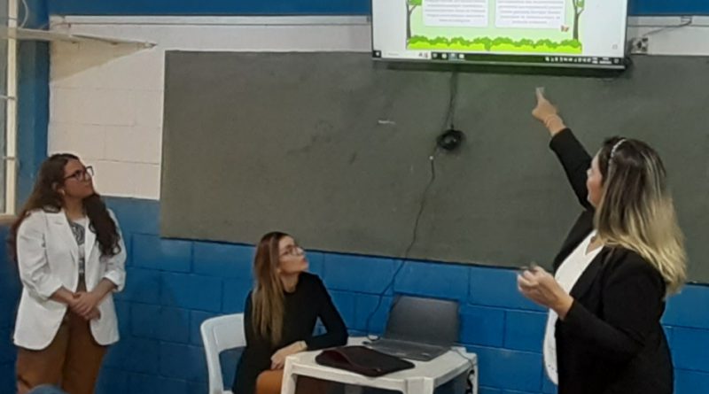 Fotografia ilustra momento da palestra. Na foto, estão três mulheres, a direita, uma delas aponta para uma televisão onde é mostrada uma apresentação. No centro, está uma mulher mais jovem, sentada passando os slides. Na esquerda, está a teceria mulher, que também gesticula.