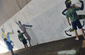 Na fotografia, adolescentes desenham sobre parede branca figuras de um jogador de futebol com a camisa da seleção, um jogador de basquete e um corredor velocista paralímpico, usando próteses no formato de lâminas.