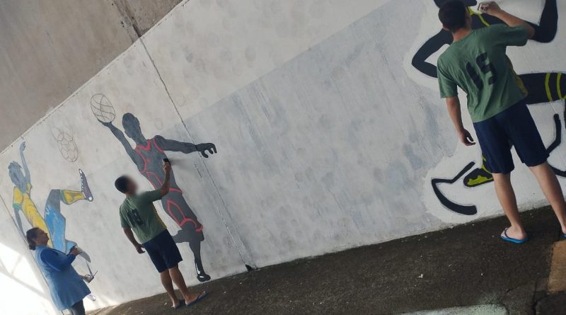 Na fotografia, adolescentes desenham sobre parede branca figuras de um jogador de futebol com a camisa da seleção, um jogador de basquete e um corredor velocista paralímpico, usando próteses no formato de lâminas.