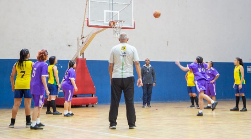 Adolescente arremessa lance livre durante partida de basquete, enquanto colegas das equipes observam ao redor da cest