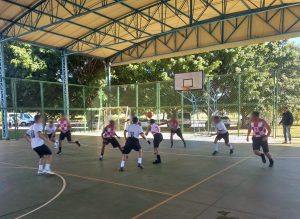 Fotografia mostra adolescentes participando de uma partida de basquete, em uma quadra coberta. O ambiente é amplo, e ao fundo, observam-se árvores e muito verde. 