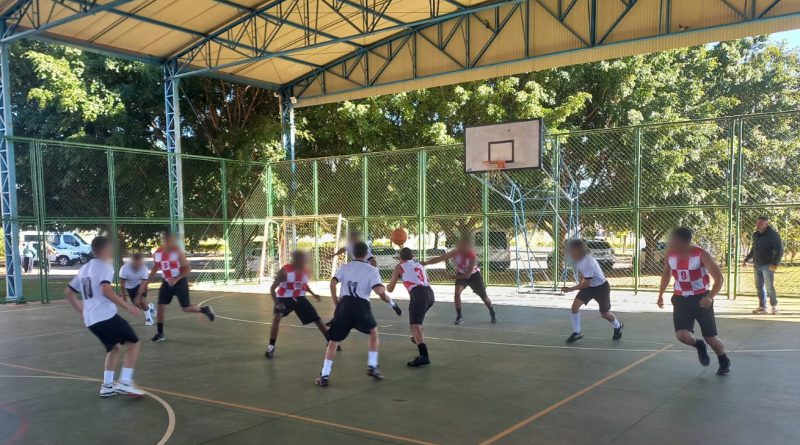 Fotografia mostra adolescentes participando de uma partida de basquete, em uma quadra coberta. O ambiente é amplo, e ao fundo, observam-se árvores e muito verde.