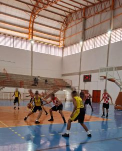 Fotografia mostra jovens disputando uma partida de basquete em um ginásio. Os jovens usam uniformes amarelos e vermelho quadriculado com branco.
