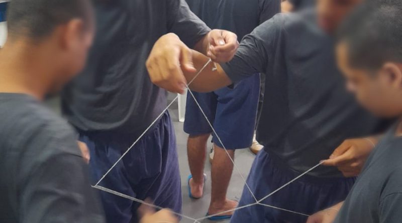 Fotografia retrata um grupo formado por quatro adolescentes que estão fazendo uma dinâmica de grupo com um fio. A dinâmica tem como objetivo gerar integração. Os jovens estão posicionados em forma de círculo e cada um deles segura uma parte do mesmo fio.