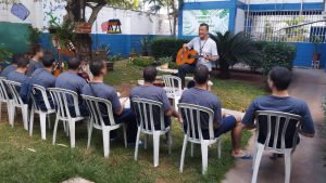 Fotografia retrata um grupo de sete jovens, todos sentados em cadeiras de plástico branco. Eles estão de costas e observam as orientações do instrutor da oficina. O instrutor usa uma camisa branca, calça escura e segura um violão. A atividade é realizada em um jardim do centro, ao lado de uma árvore.
