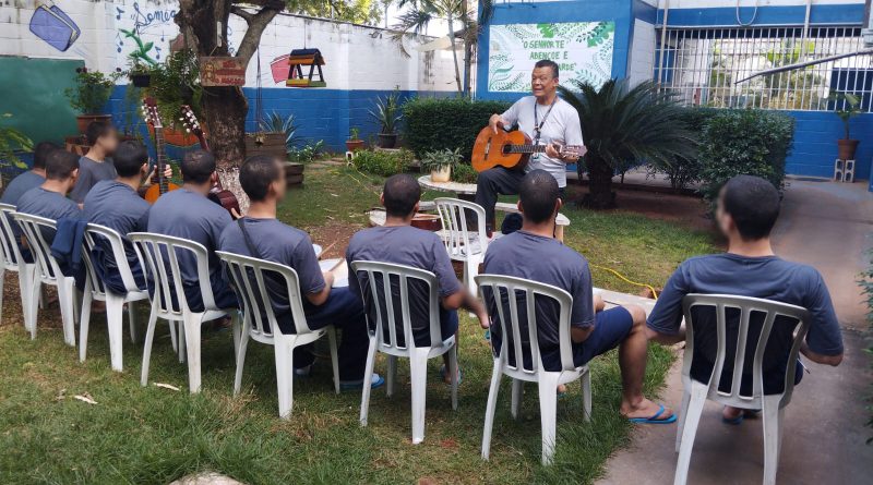 Fotografia retrata um grupo de sete jovens, todos sentados em cadeiras de plástico branco. Eles estão de costas e observam as orientações do instrutor da oficina. O instrutor usa uma camisa branca, calça escura e segura um violão. A atividade é realizada em um jardim do centro, ao lado de uma árvore.