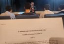 Fotografia retrata, em primeiro plano, página impressa com o conto “O Sonho de um Homem Ridículo”, do escritor russo, Fiódor Dostoiévski. Em segundo plano estão adolescentes sentados em poltronas do teatro. Ao fundo, está o juiz corregedor, Evando Pelarin