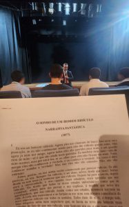 Fotografia retrata, em primeiro plano, página impressa com o conto “O Sonho de um Homem Ridículo”, do escritor russo, Fiódor Dostoiévski. Em segundo plano estão adolescentes sentados em poltronas do teatro. Ao fundo, está o juiz corregedor, Evando Pelarin