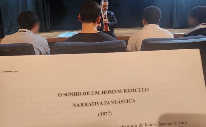 Fotografia retrata, em primeiro plano, página impressa com o conto “O Sonho de um Homem Ridículo”, do escritor russo, Fiódor Dostoiévski. Em segundo plano estão adolescentes sentados em poltronas do teatro. Ao fundo, está o juiz corregedor, Evando Pelarin