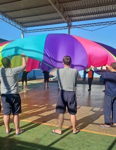 Fotografia retrata jovens balançando um pano redondo gigante colorido com as cores do arco-íris, de modo que cada jovem está na extremidade desse pano. Eles fazem o movimento de abanar, como em uma ciranda de roda. 