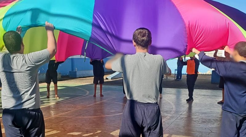 Fotografia retrata jovens balançando um pano redondo gigante colorido com as cores do arco-íris, de modo que cada jovem está na extremidade desse pano. Eles fazem o movimento de abanar, como em uma ciranda de roda.